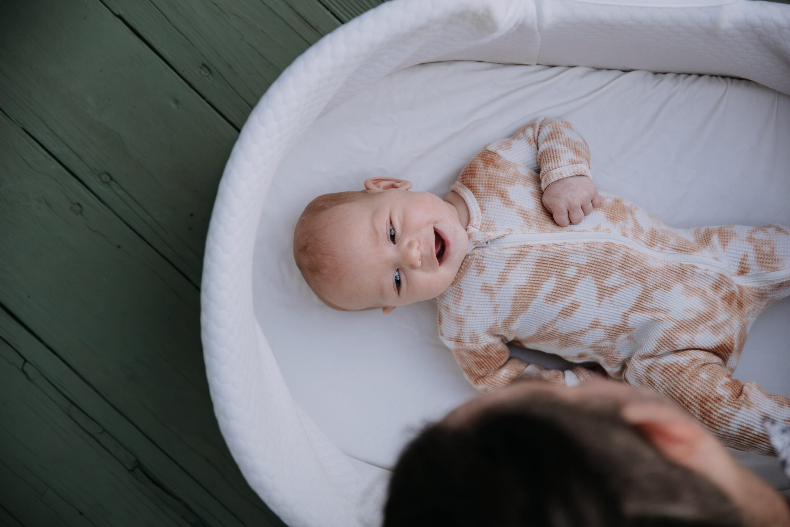 Smiling infant in Moses basket outside on porch looking at parent. 